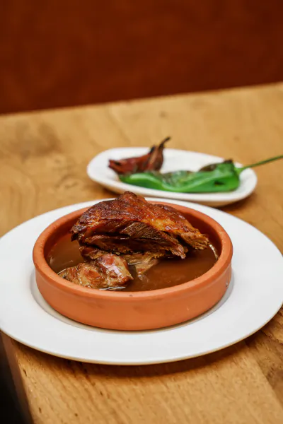 Braised meat in a terracotta bowl with rich gravy on a white plate, garnished with roasted chili peppers on a wooden table