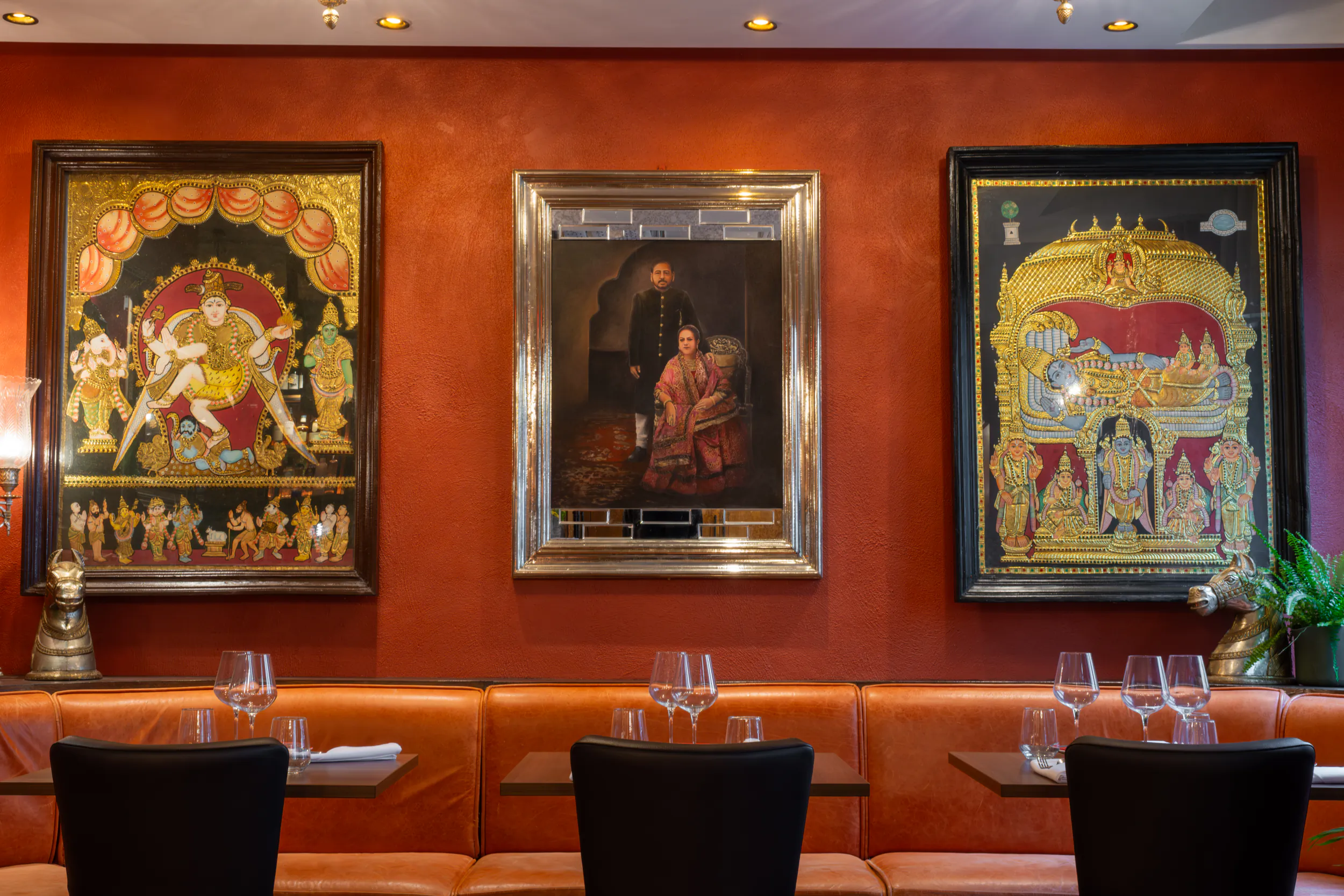 Three ornate Indian paintings on a red restaurant wall above a brown leather booth with black chairs and wine glasses