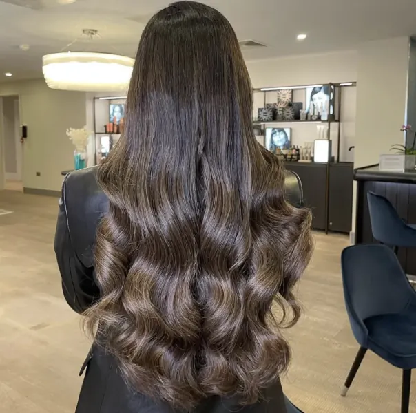 Woman with long, wavy dark brown hair viewed from behind in a modern salon with blue chairs and shelves