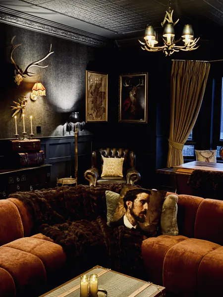 Dark luxurious lounge with orange velvet sofa, fur throw, deer antler chandelier, and framed hunting art