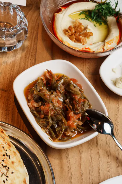 Turkish meze dishes on wooden table: bowl of hummus with walnuts and herbs, white dish of stewed greens in tomato sauce with spoon, flatbread and glass