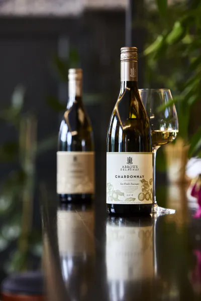 Two white wine bottles on reflective surface, front bottle labeled Abbots & Delaunay Chardonnay with wine glass