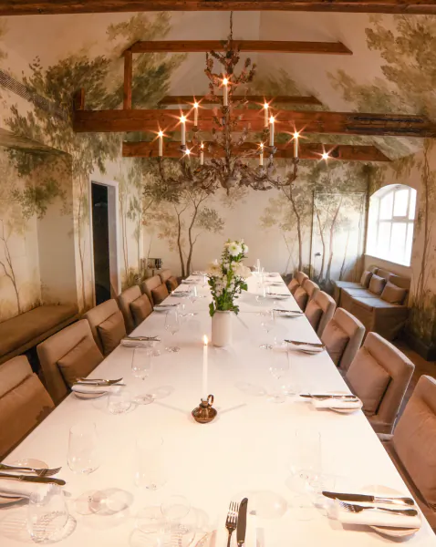 Elegant long dining table set for a formal meal in a rustic room with mural walls, lit chandelier, and white floral centerpiece