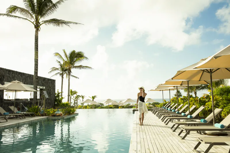 Woman in black top and white skirt walking on wooden deck beside infinity pool with lounge chairs and umbrellas at tropical resort