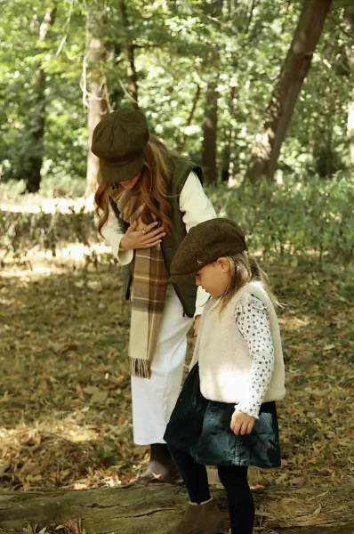 Woman and young girl in matching tweed caps and vests stand together on a forest path, woman with hand on chest