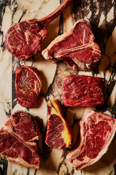 Assorted raw beef cuts including tomahawk, T-bone, and ribeye steaks arranged on a dark marble surface
