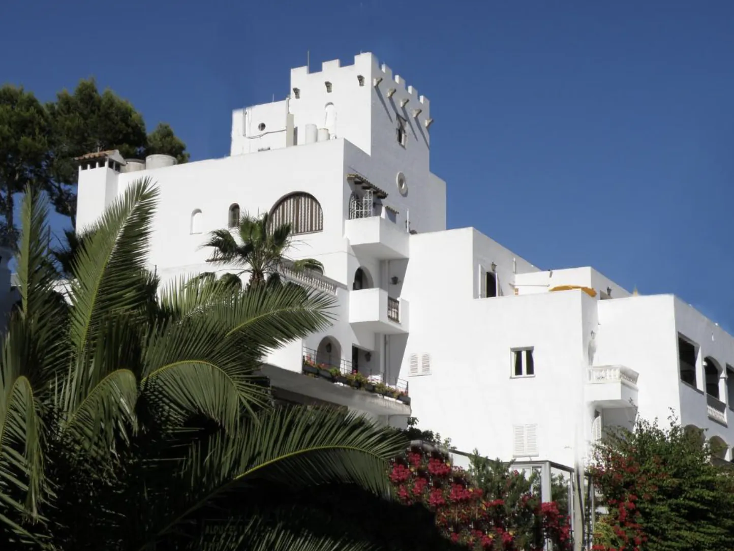 White Mediterranean-style building with castle-like tower, balconies, and palm trees under clear blue sky