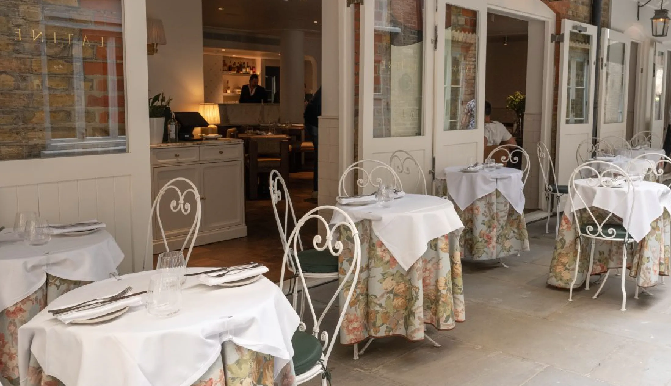Elegant restaurant patio with white-clothed tables, floral chairs, and open French doors leading to a warmly lit dining room
