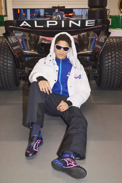 Young man in white puffer jacket and sunglasses sits in front of black Alpine F1 race car wearing matching branded sneakers