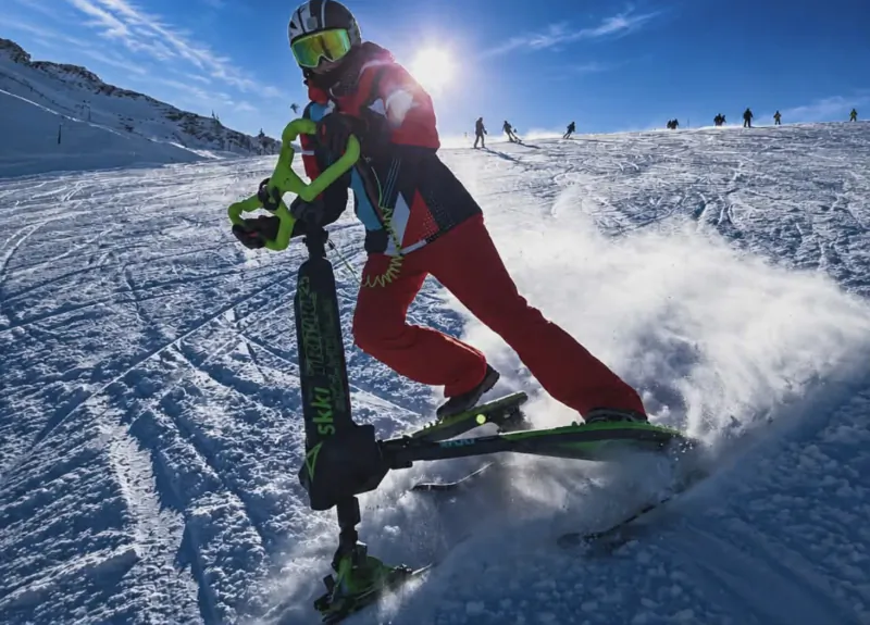Skier in red suit riding a green and black snow scooter down a snowy mountain slope with other skiers in the background