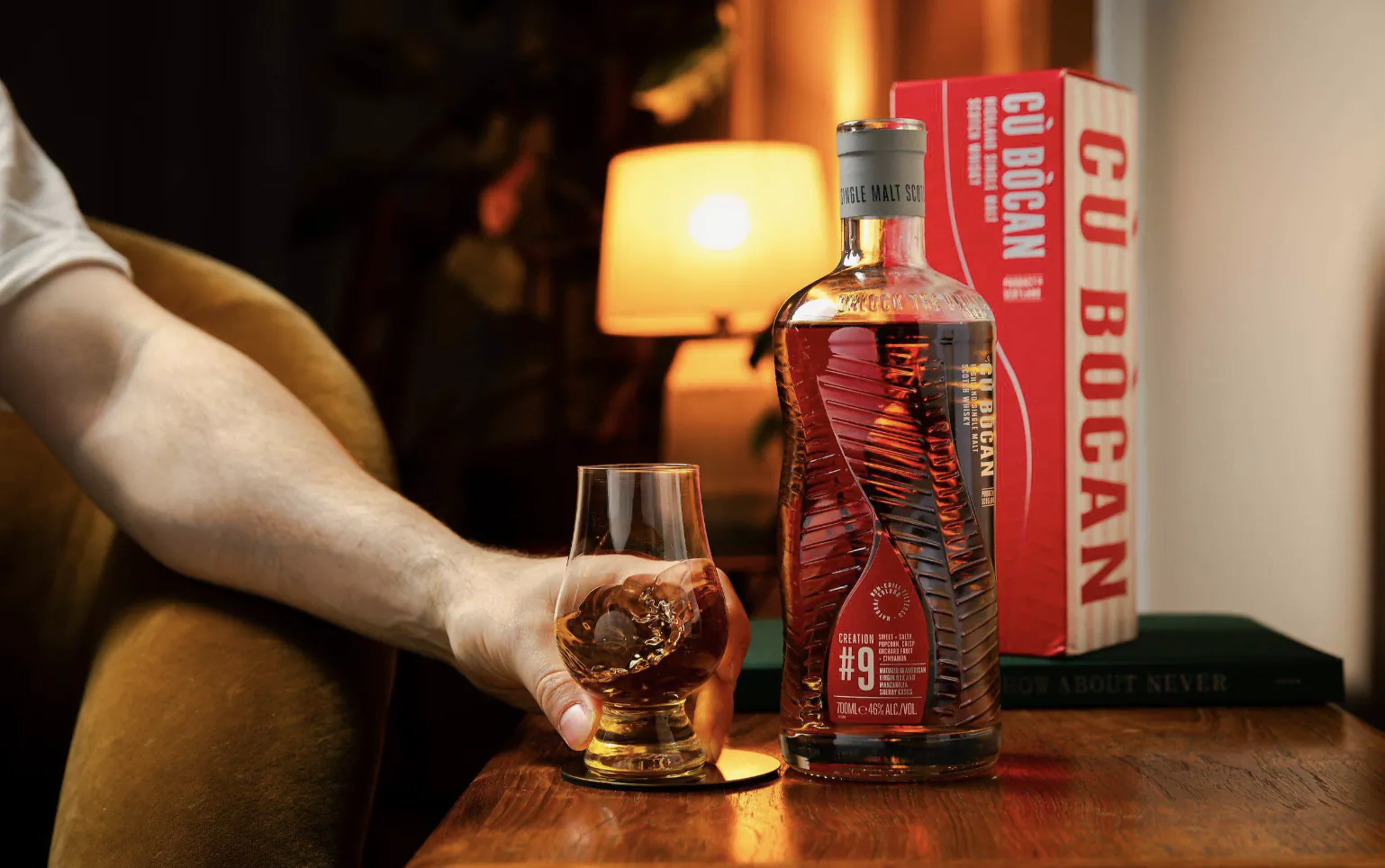 Hand holding a glass of whisky beside a Cu Bocan whisky bottle and red box on a wooden table