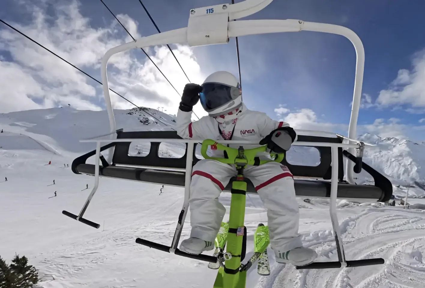 Person in white ski suit and helmet riding a green Trikke on a ski lift above snowy mountains