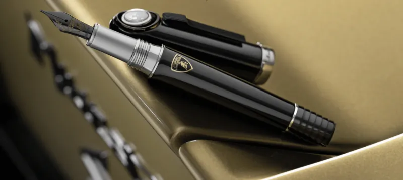 Black and silver Montegrappa fountain pen with Lamborghini logo resting on a gold surface
