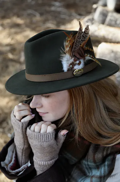 Woman in a dark green felt hat with feather detail, wearing fingerless gloves and a plaid scarf, looking down thoughtfully