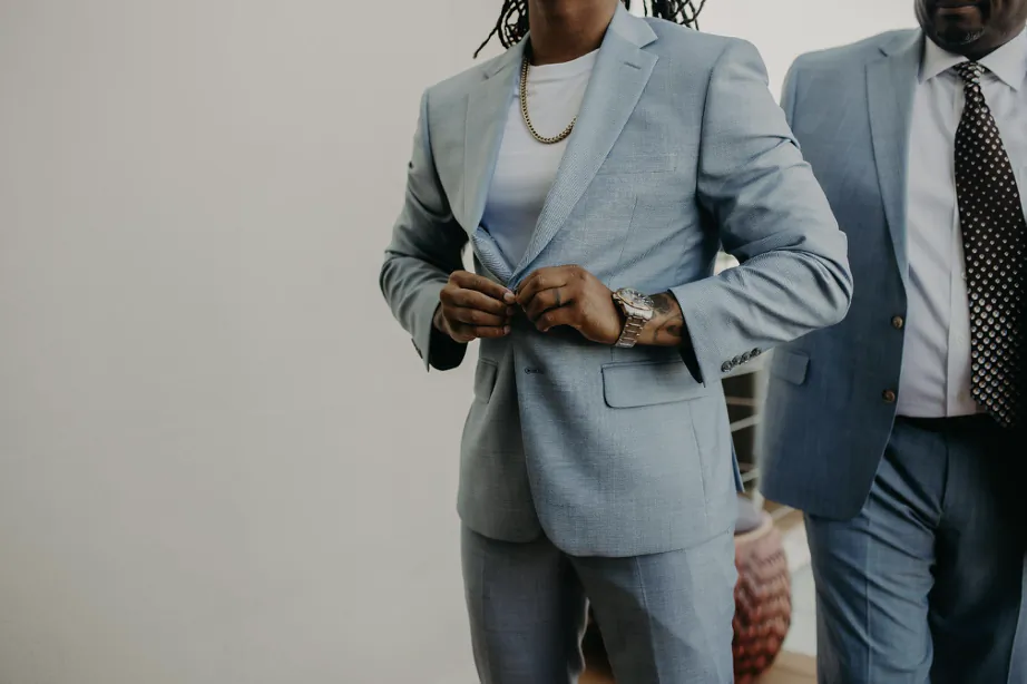 Man in light blue suit adjusting jacket buttons, another man in blue suit and polka dot tie beside him