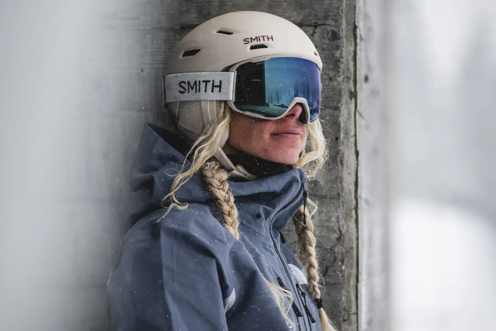 Woman in white SMITH helmet and blue goggles leaning against concrete wall, snow falling, blonde braids visible