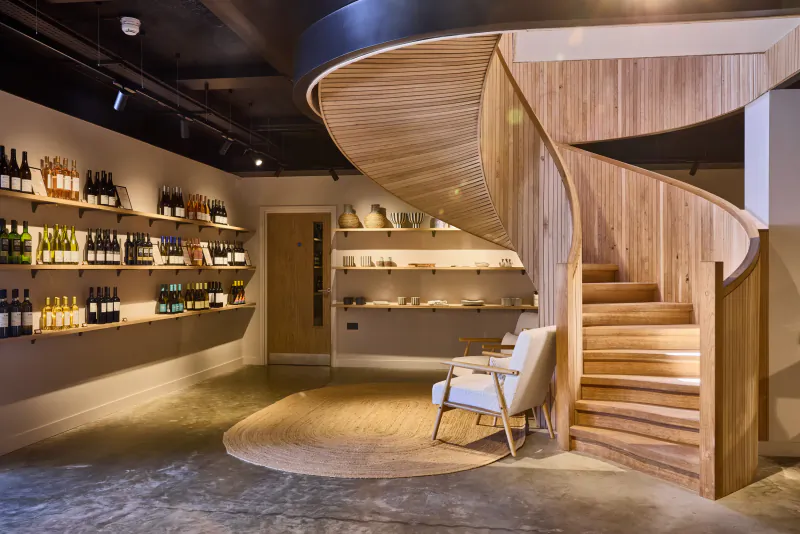 Modern wine cellar interior with curved wooden staircase, wall-mounted wine bottle shelves, white armchair on round rug, and warm lighting