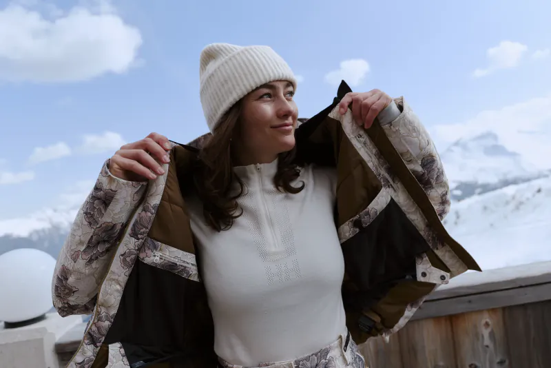 Woman in white beanie and jacket holding open her camouflage winter coat, smiling against snowy mountain backdrop