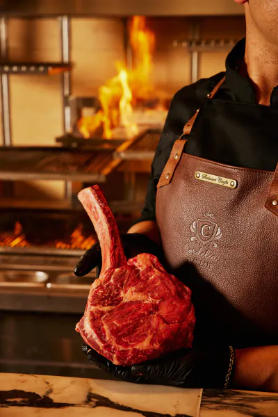 Chef in black apron holding raw tomahawk steak in front of flaming grill at Cordelia restaurant