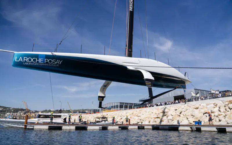La Roche-Posay AC75 racing yacht suspended by crane above water at Naples dock, with spectators on rocky shore