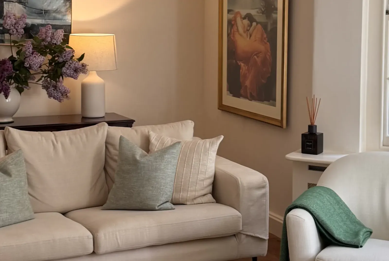 Cream sofa with sage green and beige pillows in a living room with lilac flowers, table lamp, and framed abstract art on beige walls