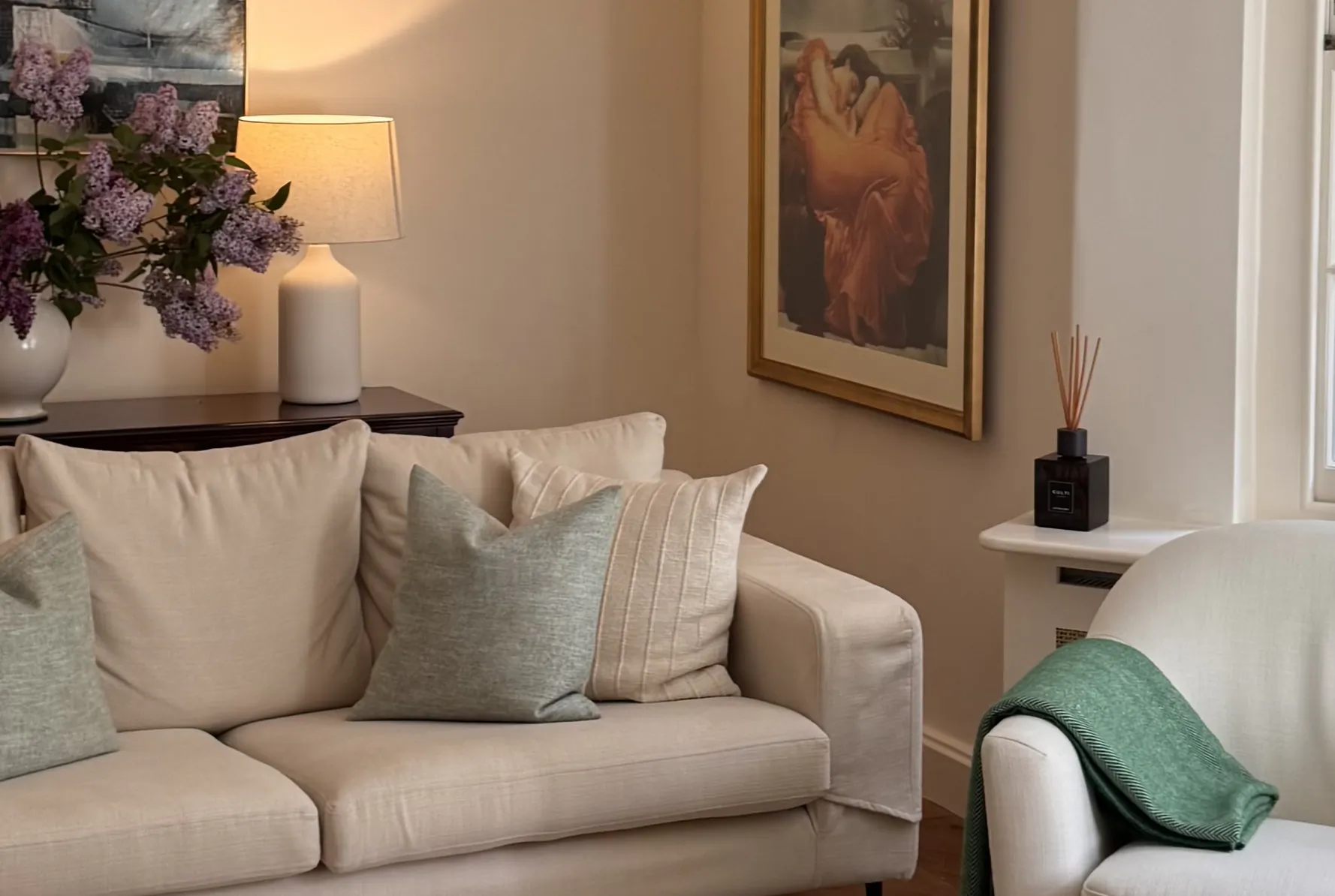 Cream sofa with sage green and beige pillows in a living room with lilac flowers, table lamp, and framed abstract art on beige walls