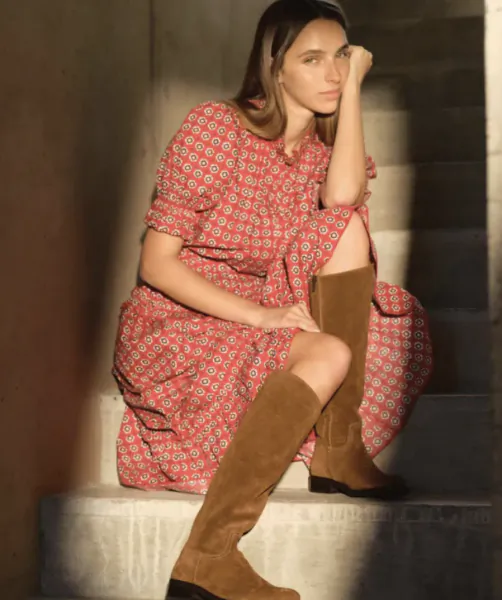 Woman in red patterned dress and brown suede knee-high boots sitting on stone steps, hand resting on cheek