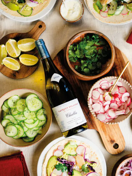 Overhead view of a festive table with a bottle of Laurent Miquel Chablis wine surrounded by bowls of sliced cucumbers, radishes, cilantro, and lime wedges on wooden boards