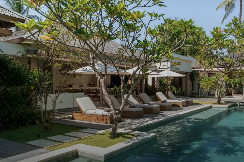 Luxury resort poolside with wooden lounge chairs, umbrellas, and tropical trees overlooking a swimming pool