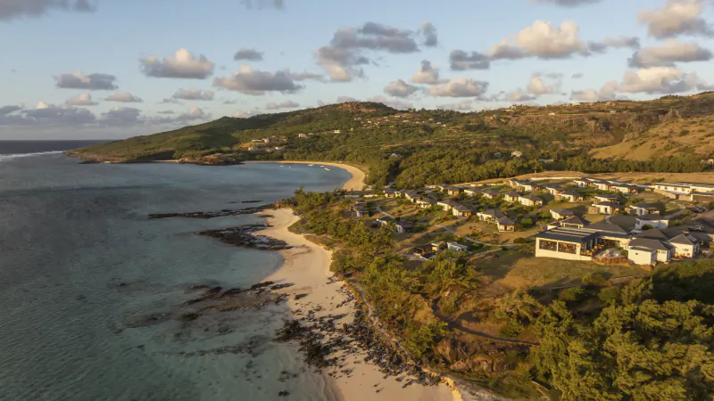 Aerial view of Constance Tekoma resort in Rodrigues, Mauritius, showing white villas nestled among green hills above a sandy beach and turquoise ocean at sunset