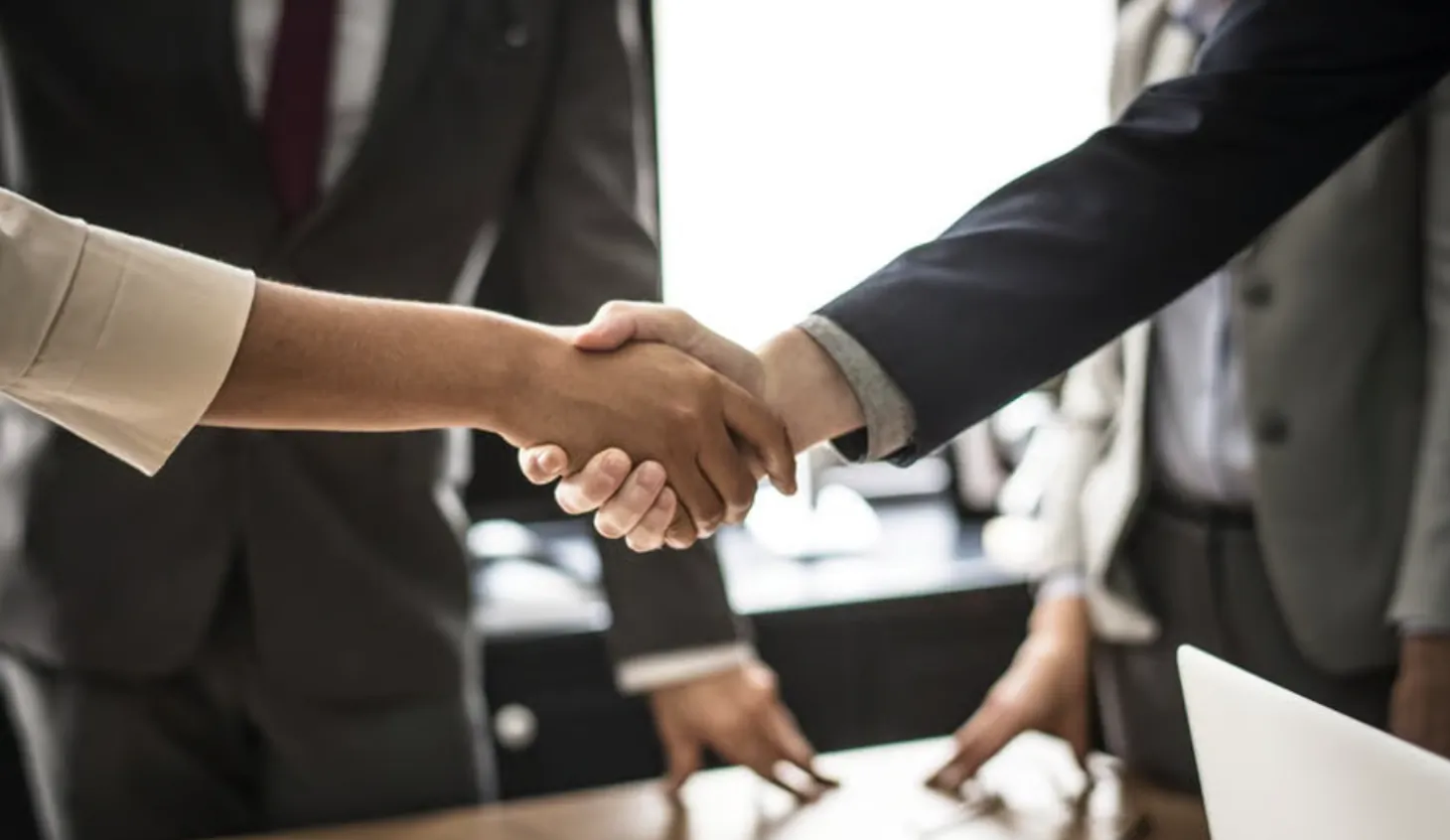 Business professionals in suits shaking hands over a conference table with a laptop nearby