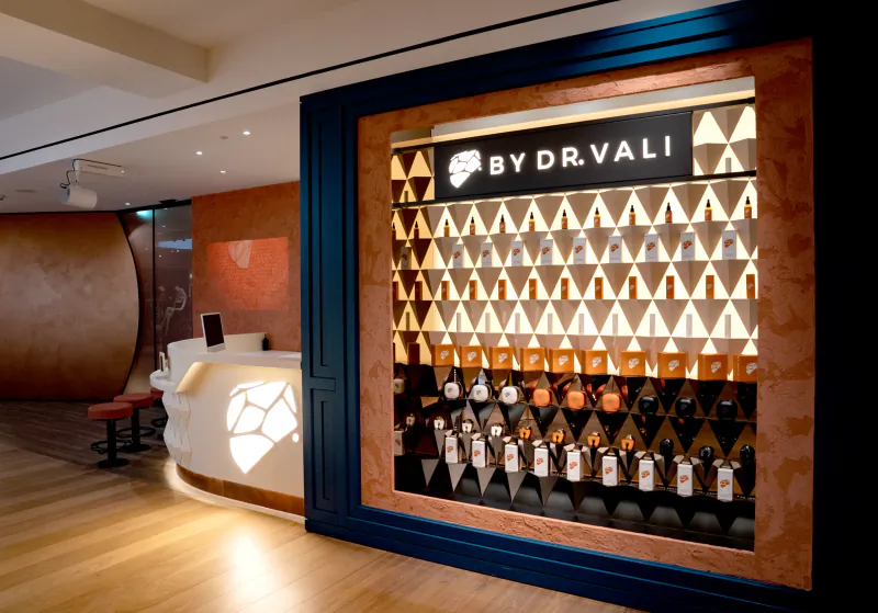 Modern skincare store display wall with illuminated bottles arranged in geometric pattern, branded 'BY DR. VALI' with lion logo
