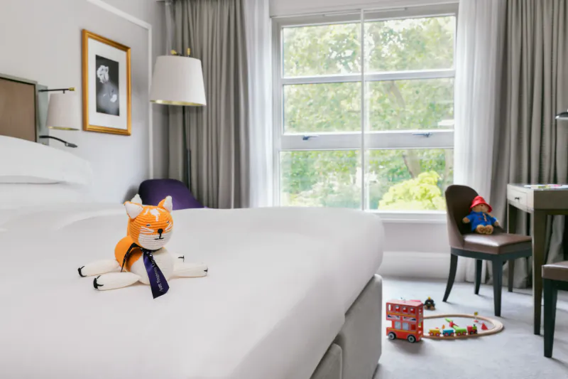 Modern hotel bedroom with white bed, orange fox plush toy, children's toys, large window overlooking trees