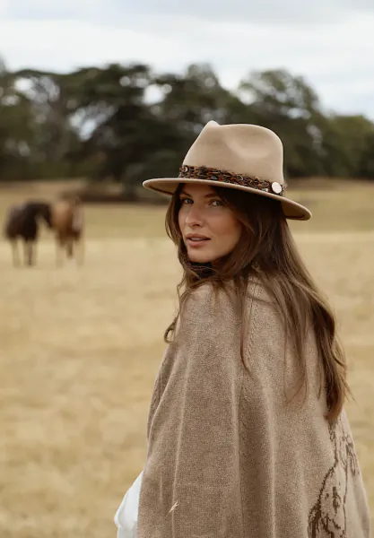 Woman in beige felt hat and wool poncho standing in a field with horses in the background