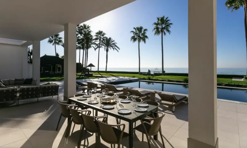 Luxury outdoor dining table set for eight on a modern villa terrace overlooking an infinity pool, palm trees, and the ocean at sunset