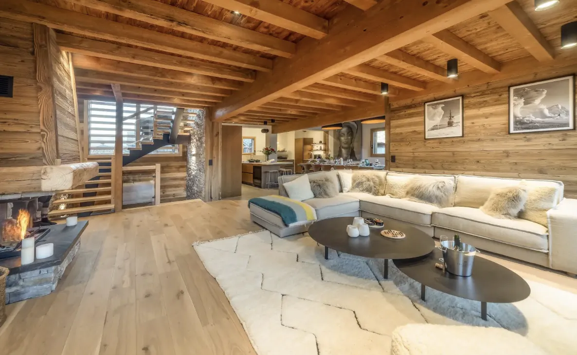 Modern open-plan alpine living room with wooden beams, stone fireplace, large L-shaped sofa, and staircase in a luxury chalet