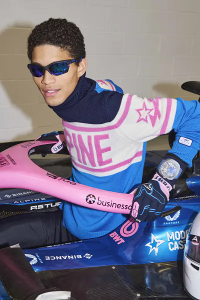 Young man in blue and pink Alpine racing sweater holding pink steering wheel, wearing sunglasses and gloves with sponsor logos