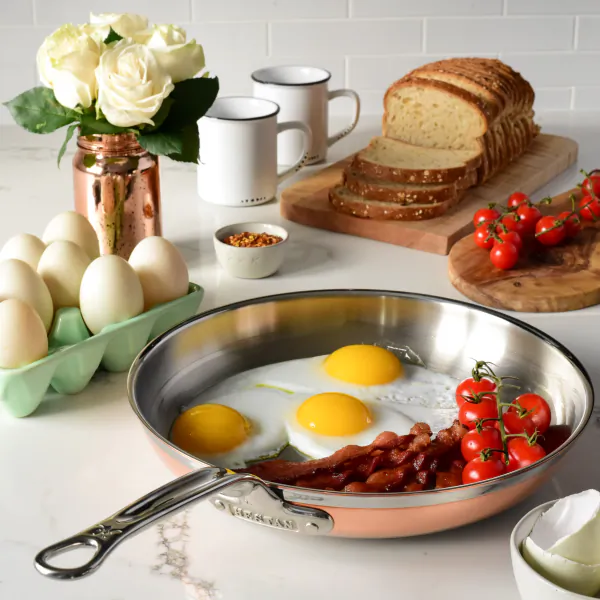 Hestan copper frying pan with sunny-side-up eggs, bacon strips, and cherry tomatoes on a white marble countertop with eggs, bread, and white roses