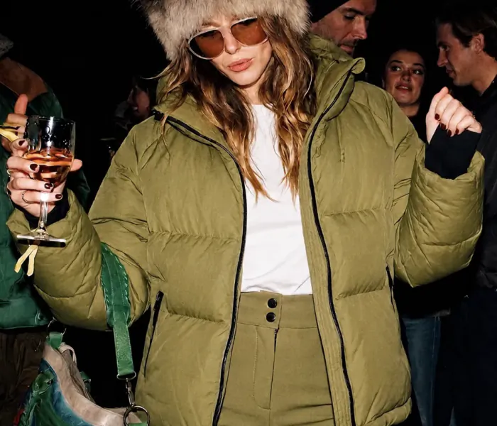 Woman in green puffer jacket and fur hat holding a cocktail at a crowded party