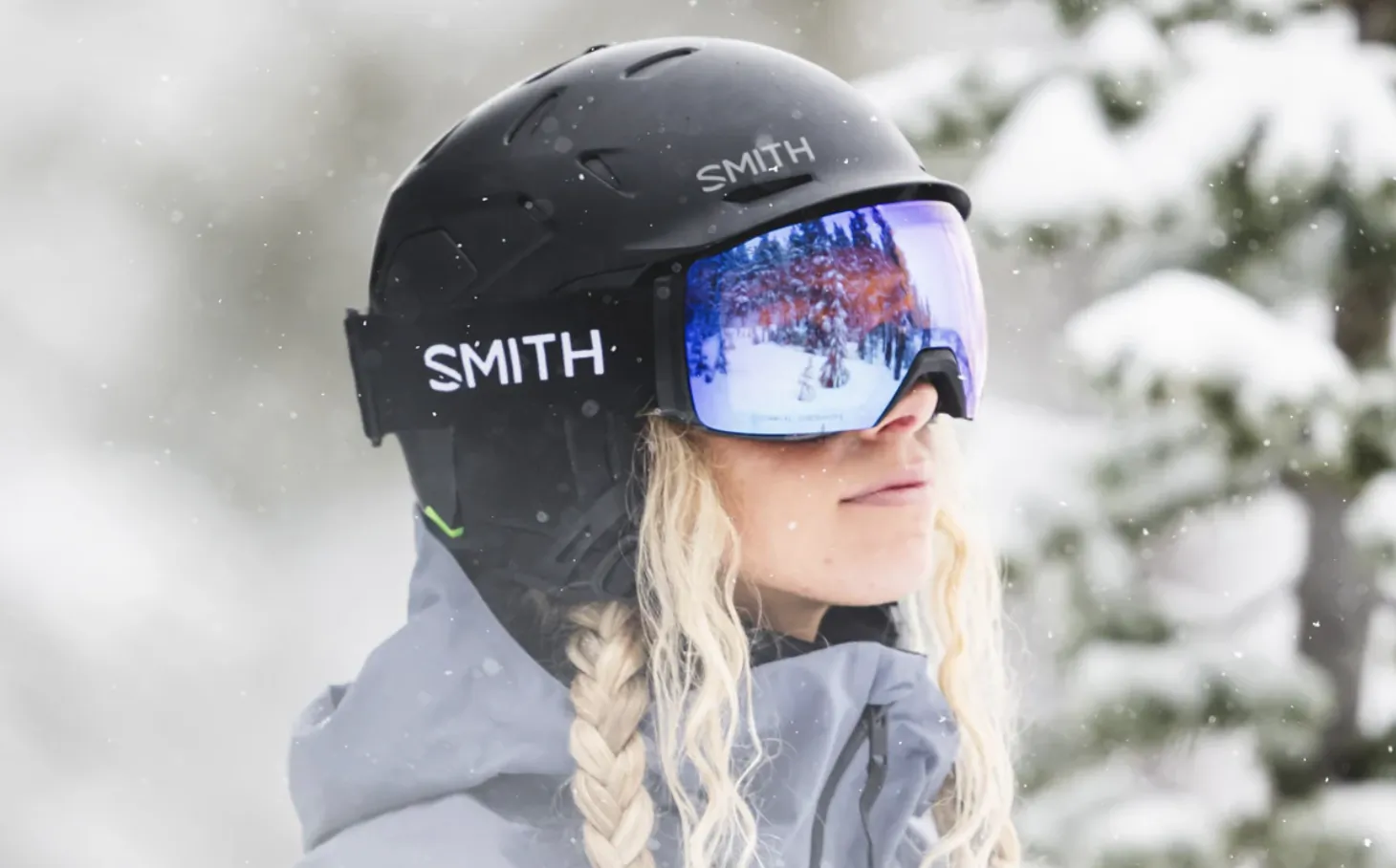 Woman in black Smith ski helmet and reflective goggles with blonde braid, snowy forest background