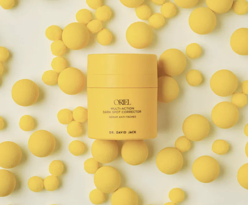Yellow Oriel Multi-Action Dark Spot Corrector serum jar surrounded by scattered yellow spheres on white background