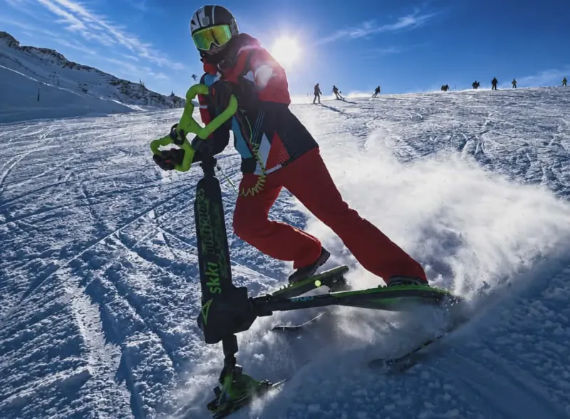 Skier in red suit riding a green Skki Trikke snow scooter down a snowy mountain slope, kicking up powder under bright sun