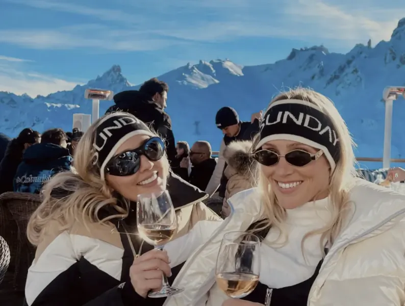 Two smiling blonde women in winter jackets and 'SUNDAY' headbands toast with wine glasses on a snowy mountain terrace
