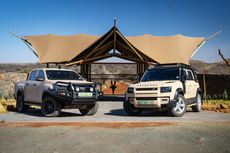 Two beige Toyota pickup trucks and SUVs parked in front of a modern desert pavilion with a large beige canopy and stone wall backdrop