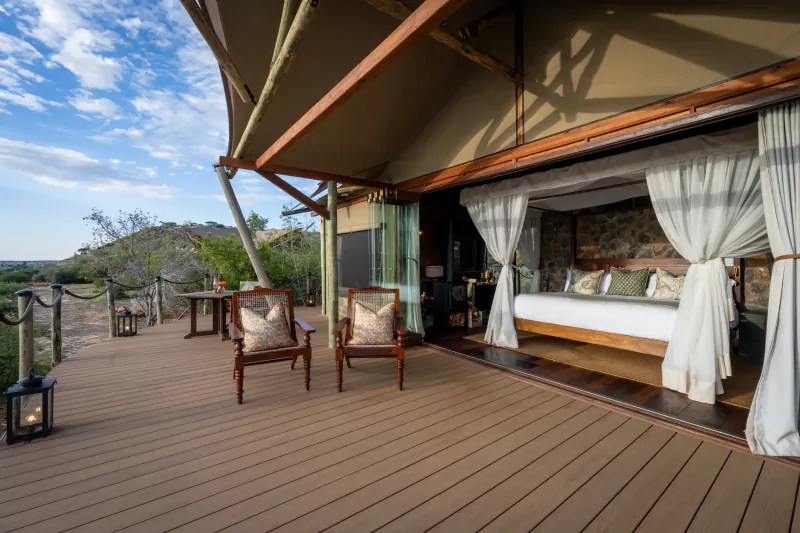 Luxury safari tent suite with wooden deck, two patterned armchairs, and open bedroom with white linens overlooking scenic landscape
