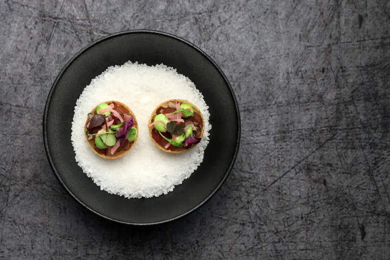 Two small round appetizers with avocado and microgreens on a bed of white salt in a black bowl