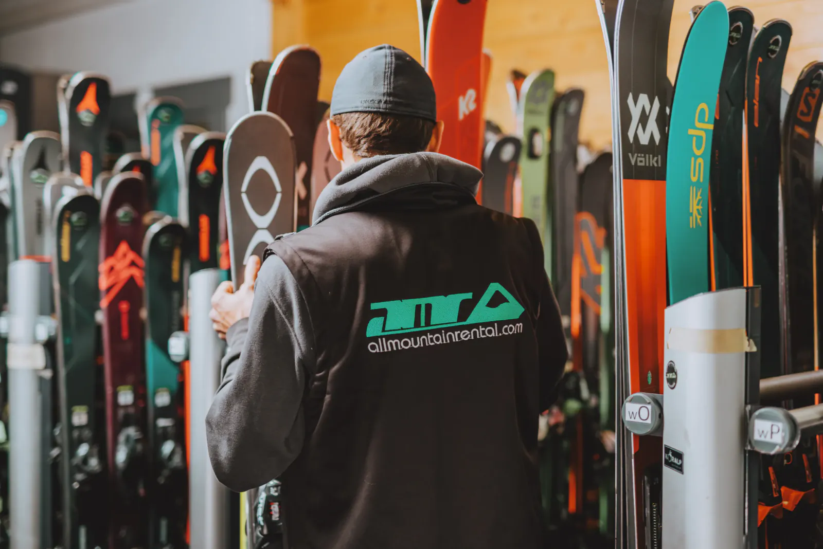Man in All Mountain Rental jacket holding skis in a ski shop surrounded by colorful rental skis