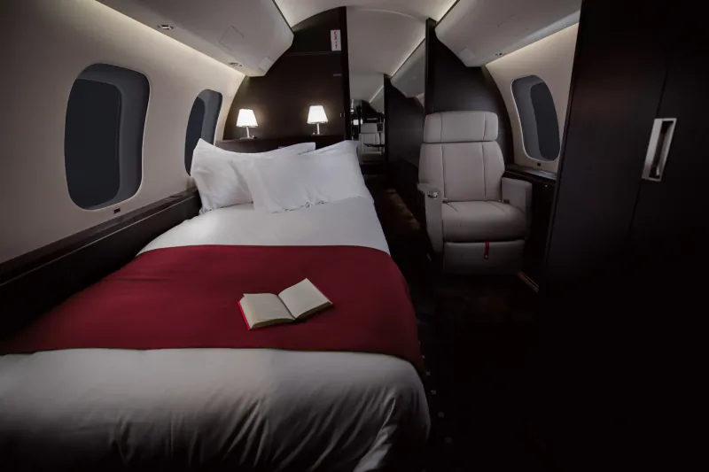 Luxury private jet bedroom with a neatly made bed, red throw, open book, and cream leather seat beside oval windows