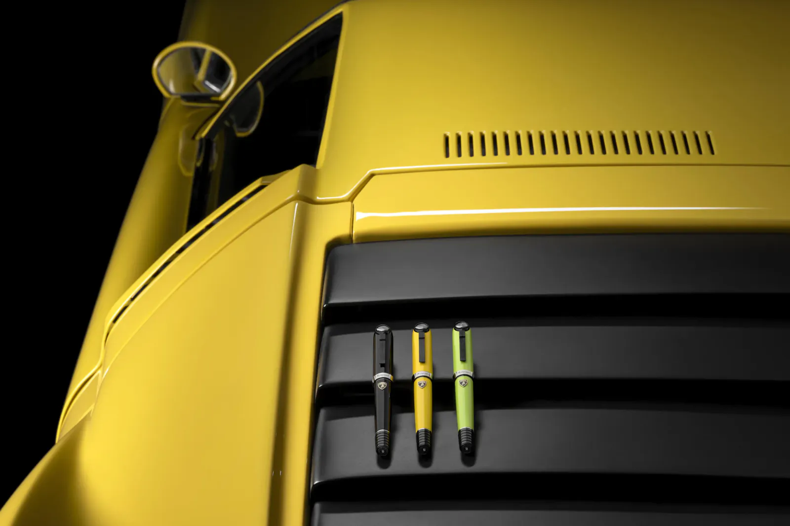 Three luxury pens in black, yellow, and lime green rest on the black air intake of a yellow Lamborghini sports car