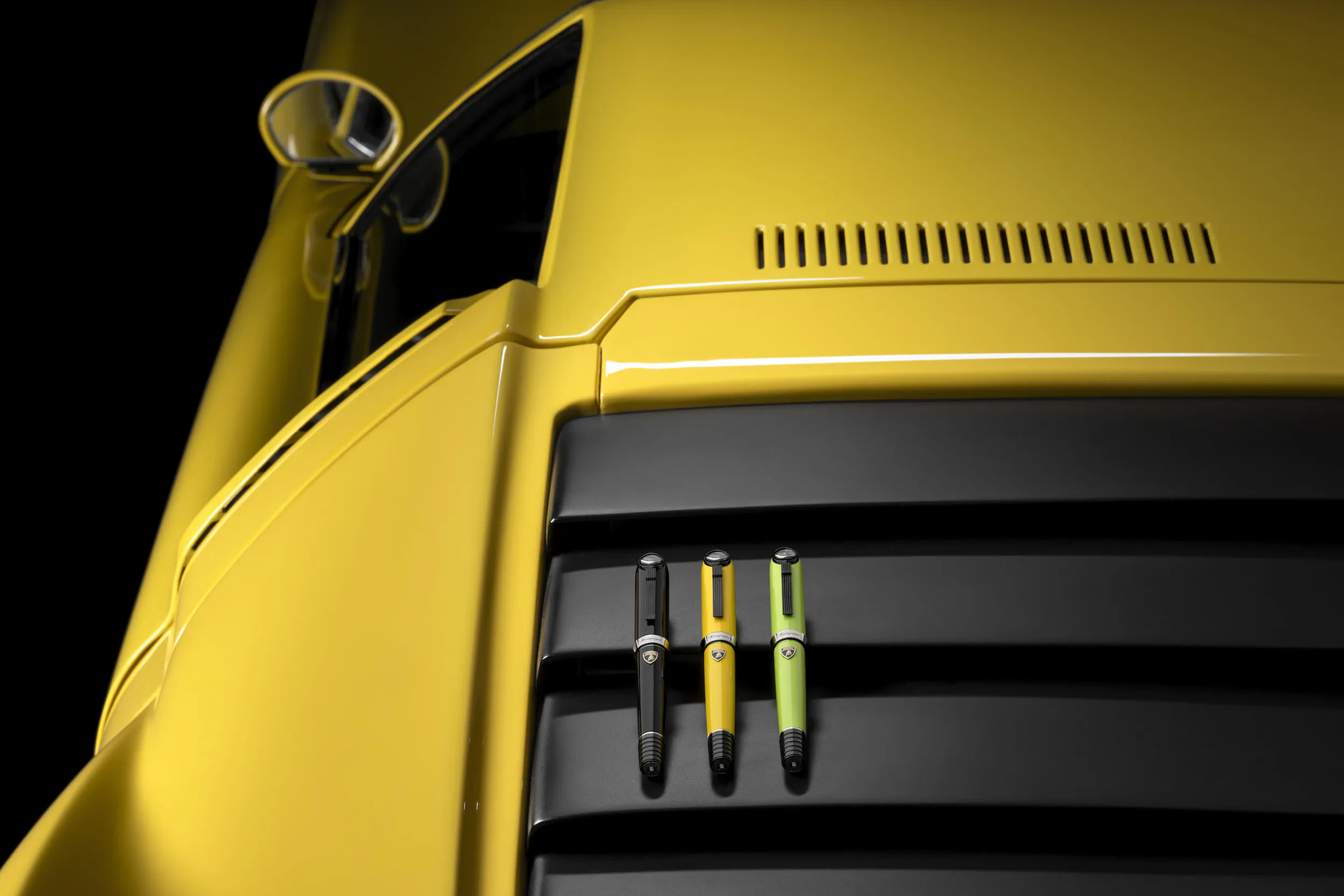 Three luxury pens in black, yellow, and lime green rest on the black air intake of a yellow Lamborghini sports car